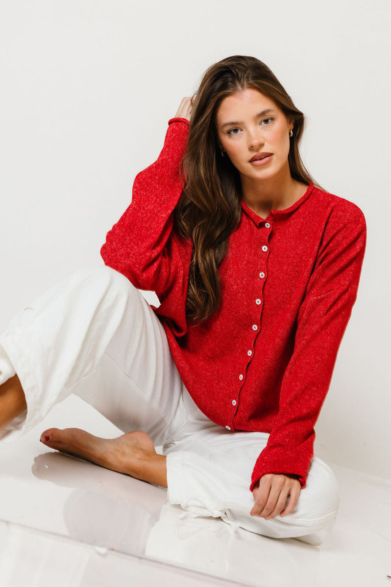 Model seated on the floor in the Piper button-front cardigan in bright scarlet red, worn with white pants