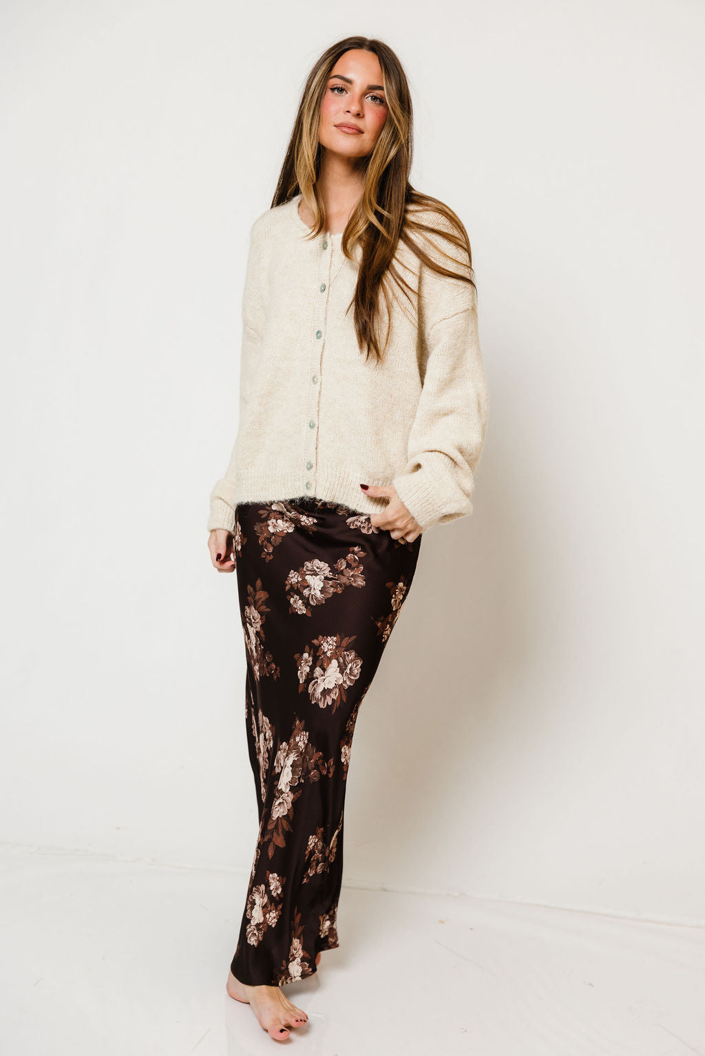 Model in the Kirana satin maxi skirt in dark brown with a peach floral print, worn with a cream knit sweater