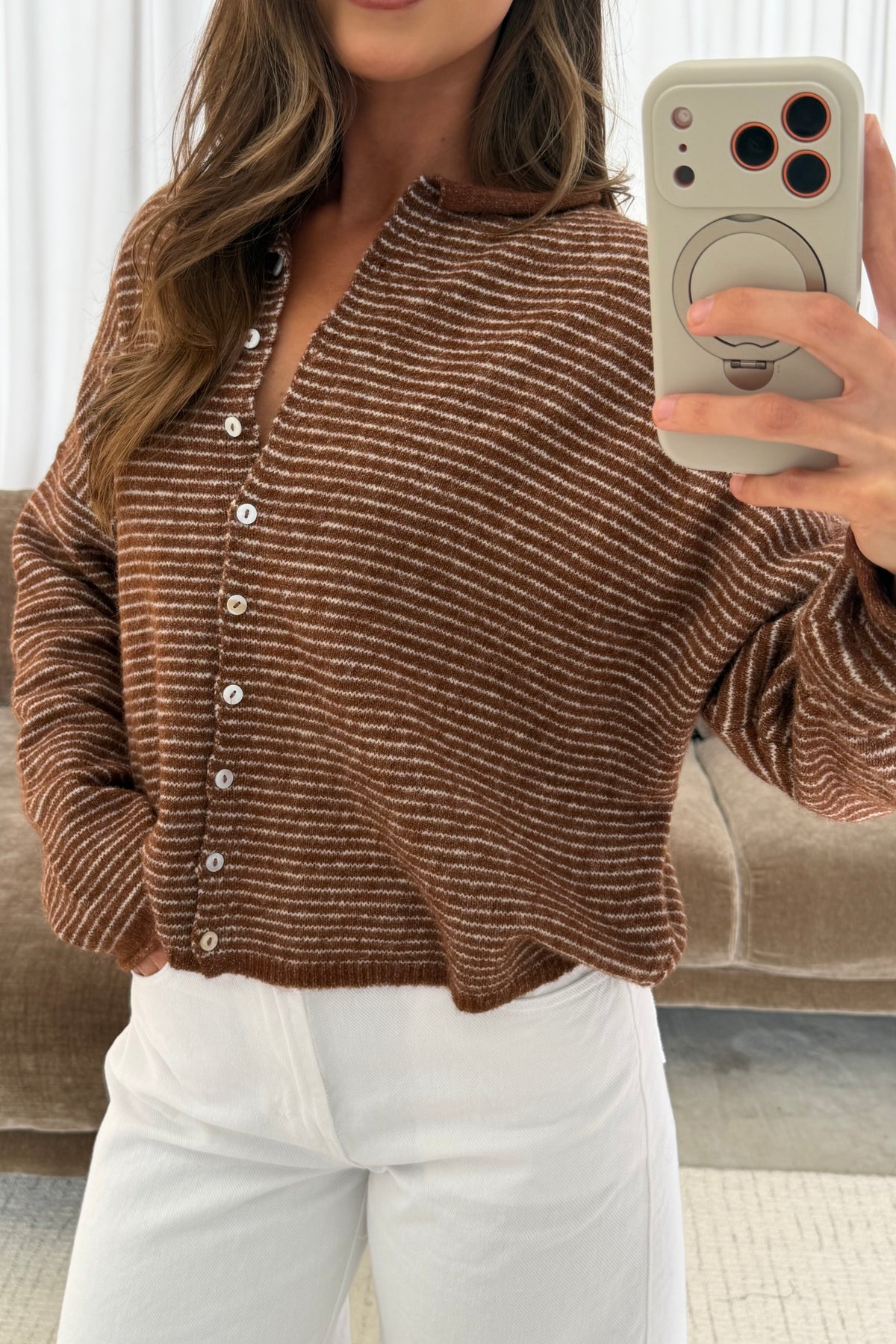Mirror selfie of a model in the Piper button-front cardigan in brown with thin cream pinstripes, worn with white pants