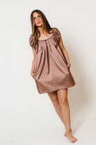 Model in the Ana satin mini dress in mocha with short puff sleeves, a gathered square neckline and pockets
