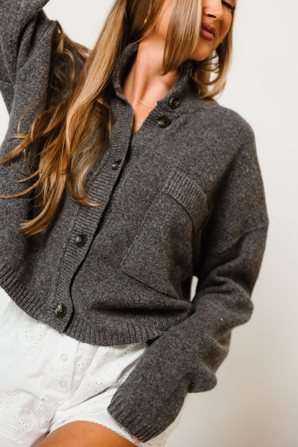 Model in the Winslow funnel neck cropped knit cardigan in dark charcoal grey with a button front and chest pocket, worn with white shorts