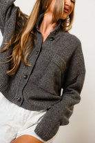 Model in the Winslow funnel neck cropped knit cardigan in dark charcoal grey with a button front and chest pocket, worn with white shorts