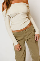Model in the Twyla ribbed knit off-the-shoulder sweater in cream with long sleeves, worn with olive pants