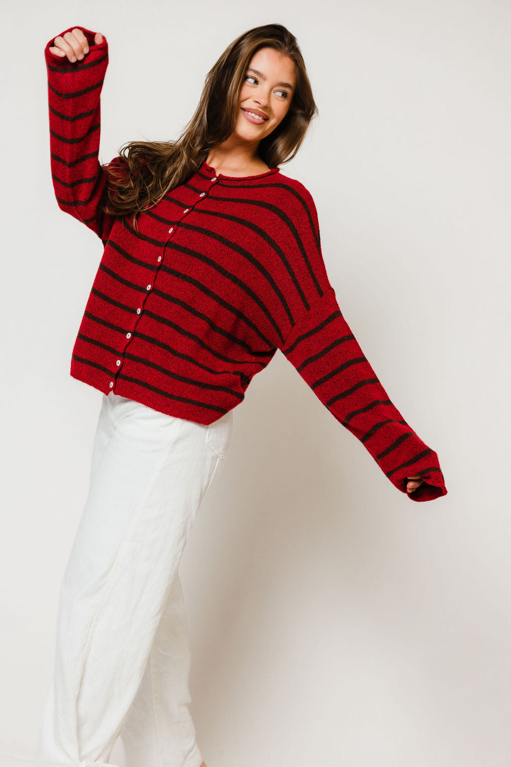 Model with one arm raised in the Piper button-front cardigan in red with thin dark brown cabana stripes, worn with white pants