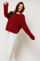 Model with one arm raised in the Piper button-front cardigan in red with thin dark brown cabana stripes, worn with white pants